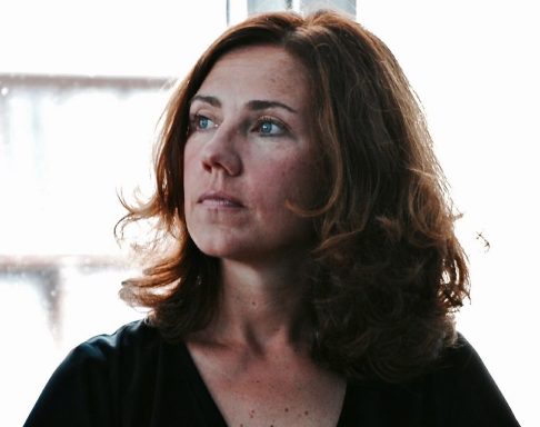 Frau mit lockigem, braunem Haar, nachdenklichem Ausdruck und direktem Blick.